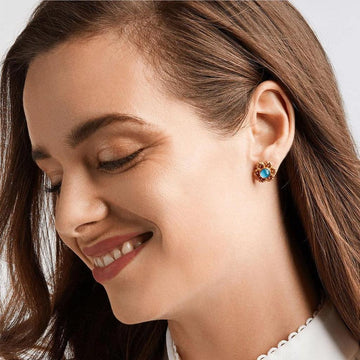 Gold stud earrings with a blue gemstone center, shown on a model's ear.
