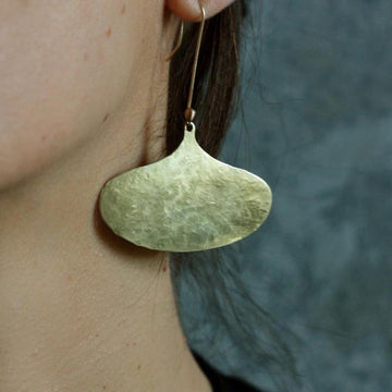 Brass teardrop earrings with hammered texture, worn against a dark background.
