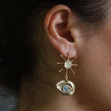 Gold earrings featuring a sunburst design and a blue eye charm, worn against dark hair.
