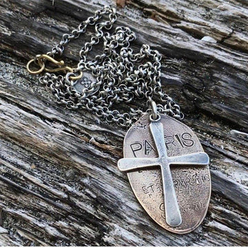 Silver chain necklace with an oval bronze pendant featuring a cross and engraved lettering, displayed on wood.
